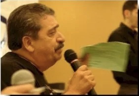 A man with dark hair and a mustache speaks into a microphone while holding a green index card.