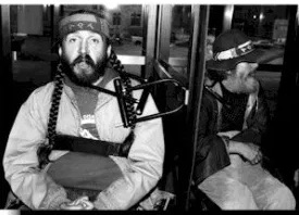 Black and white picture of a man with a full beard and mustache in a hat. What appears to be a bike lock hooked to a pole and a strap on his arm.