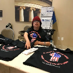 Person in a red cap and dark tee shirt sits behind a table displaying similar dark tee shirts.
