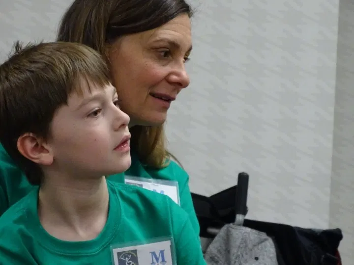 A woman with dark hair sits behind a young boy with dark hair. Both wear a green shirt with nametags