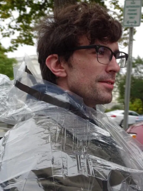 Photo of Liam Dougherty Man with dark hair and eye glasses wearing a rain poncho