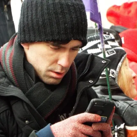 Picture of Liam Dougherty Person with a black cap, dark scarf and coat peering into a cellular phone screen.