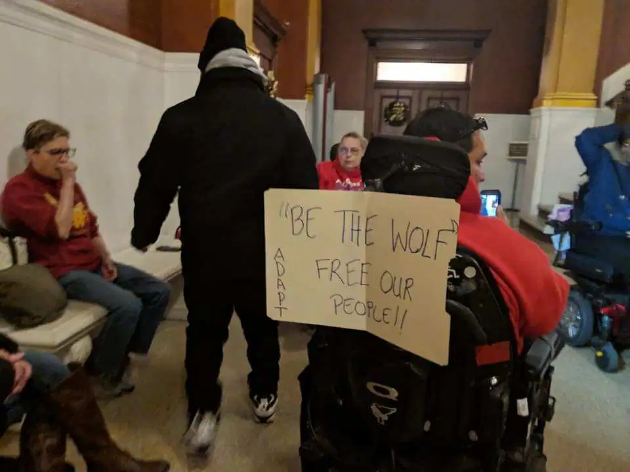 People are spread out in a hallway. Foreground: The back of a person in a power wheelchair with a sign saying "Be The Wolf Free Our People ADAPT"