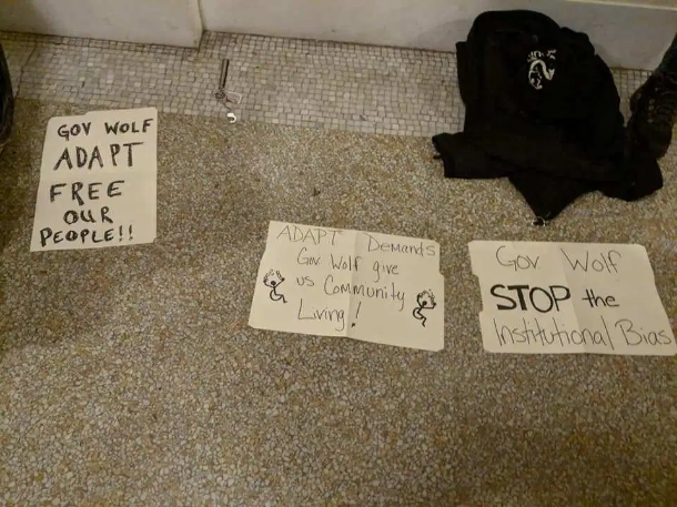 Three signs spread out on a floor. "Gov Wolf ADAPT Free Our People!!, "ADAPT Demands Gov Wolf give us Community Living", and "Gov Wolf STOP the Institutional Bias."