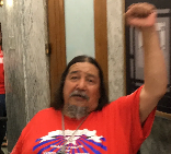 Picture of Ed Sandoval A person with long dark hair and a red shirt raises the power fist.