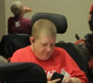 Cal Montgomery Person in a power wheelchair, buzz cut, and red tee shirt looking at a cell phone.