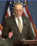 A man in a dark suit, white shirt and light tie stands at a podium speaking into a microphone.