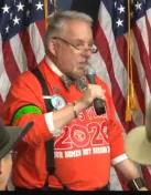 A man in a red shirt and suspenders speaks into a microphone.