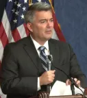 A man in a dark suit speaks into a microphone. He is holding a piece of paper in his free hand.