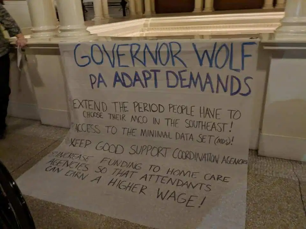 A sign "Governor Wolf PA ADAPT Demands
Extend the period people have to choose their MCO in the Southeast!
Access to the minimal data sets (MCO)!
Keep good support coordination agencies!
Dramatically increase funding to home care agencies so that attendants can earn a higher wage!"
