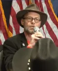 A man speaks into a microphone in front of an American flag