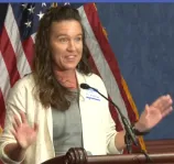 A woman with long, dark hair and a light suit speaks into a microphone.