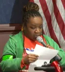 A Black woman with braids, wearing a green sweater holds a sheet of paper and speaks into a microphone.