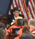 A person in a cowboy hat and dark suit speaks in front of a crowd.