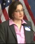 Woman in a dark jacket and pink shirt speaks into a microphone.