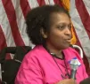 A Black woman in in a wheelchair and a pink top speaks into a microphone. 