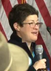 A woman with short cut hair and a dark top speaks into a microphone.