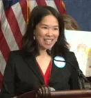 A woman with dark hair and a dark suit speaks into a microphone. An American flag is in the background.