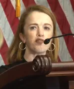 A woman with long hair, gold hoop earrings stands behind a podium and speaks into a microphone.