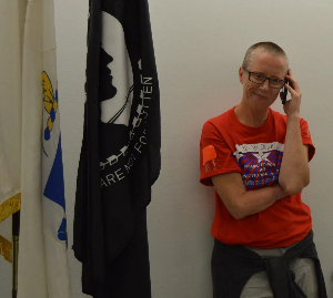 Person with a buzz cut, red shirt and blue pants and glasses holds a cellphone to their face and stands next to a black flag