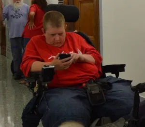 Picture of a person in a black power wheelchair wearing a red shirt and dark pants, looking at a cell phone.