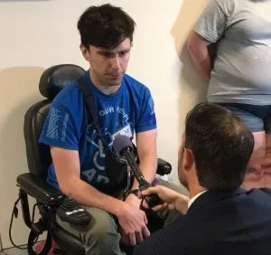 A person in a blue shirt and dark hair sits in a power wheelchair. They are speaking to a man in a dark suit holding a microphone.