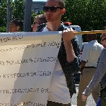 A person with dark hair and sunglasses holds up a handwritten sign.