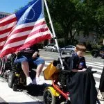 Picture of a young person in a power wheelchair carrying a modified American Flag. The stars are in the form of a wheelchair.