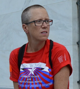 Carol, an ADAPT activist from Washington DC Person with a buzz cut, glasses and a red shirt peers off into the distance.