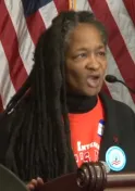 A Black woman with long dreadlocks wearing a red shirt and dark jacket speaks into a microphone. An American flag is in the background.