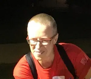 Picture of Carol Tyson A person with a short buzz cut, glasses and a red top looks into the distance.