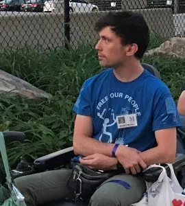 Picture of Liam Dougherty A man in a blue tee shirt and dark pants sits in a wheelchair and stares off into the distance.