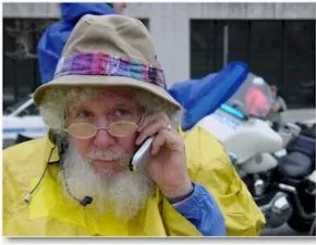A man with a white beard, tan hat and yellow top speaks into a cellphone while looking away from the camera