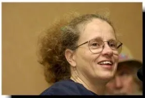 Woman with a curly ponytail and eye glasses smiles as she looks into the distance.
