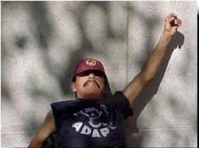 A man in a red cap and dark "ADAPT" sleeveless top raises his fist in protest.