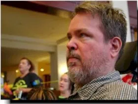 A man with a beard and mustache with a button-down shirt sits in a room with other people, watching something off in the distance.