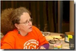 Picture of Stephanie Thomase of ADAPT Woman with curly hair in a ponytail and glasses, wearing a red top speaking to someone off camera