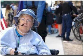 A woman in a light blue top and blue hat, with an earpiece around her ears sits in a dark power wheelchair