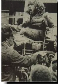 Black and white photo of a protestor in a wheelchair. He appears to be shouting or chanting. There are two men standing in the foreground.