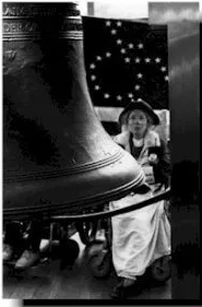 Black and white photo of a giant bell in the foreground. A woman in a hat is positioned in front of an American flag. The flag's starts are in the shape of a wheelchair.