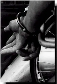 Black and white image of two hands crossed and handcuffed behind a wheelchair. The wheels are seen on the right of the frame.