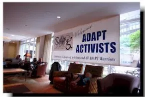 Picture of a large room with wall to ceiling windows. A large sign that says "ADAPT ACTIVISTS" hangs across the windows.