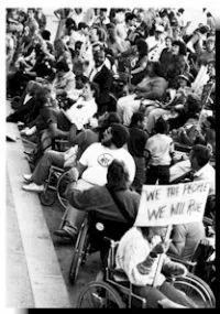 A Black and white photo of a large crowd, some in wheelchairs and some standing, gathered outdoors. 