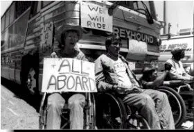Black and white image of protestors in wheelchairs blocking a bus.  A sign on the front of the bus says "We will ride".  One person holds a sign that says "All Aboard".