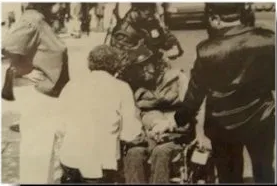 Black and white image of ADAPT protestor in a wheelchair. There are three people standing around the protestor. One is kneeling in front of the protestor.