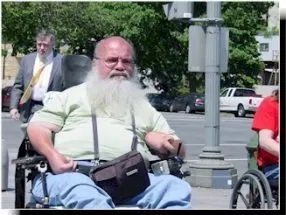 A man with a white beard, green top, and blue jeans sits in a power wheelchair during a protest.