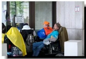 A man in a blue top and rain poncho sits in a wheelchair behind other wheelchair users wearing rain ponchos. One holds a sign.