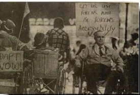 Sepia-toned image of protestors in wheelchairs and handwritten signs demanding accessibility