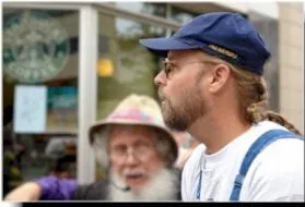A man in the foreground with a blue cap and white top looking to the left.  A man in the background with a white bushy beard and bucket hat looks towards the foreground.