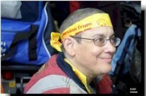 Picture of Spitfire Sabel A person with a shaved head wears a yellow head band with red writing and eyeglasses. They have a smile on their face.